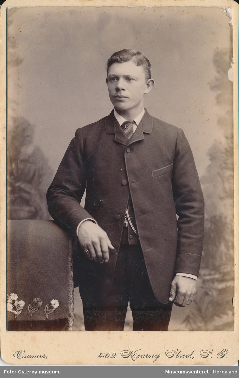 atelierfotografi av ung mann med mørk dress med vest, skjorte med striper og kvit krage som står og lener eine armen mot ein stolrygg