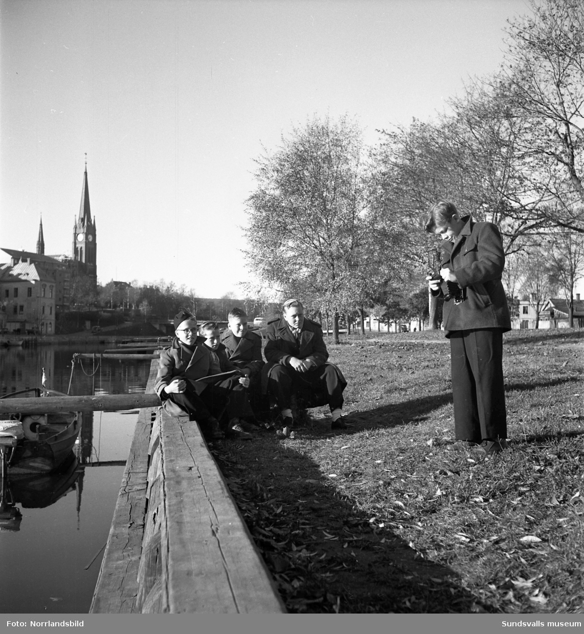 Stockholms gosskör på Sundsvallsbesök. Fyra pojkar poserar vid Selångersåns norra kajkant och en femte pojke står vid sidan om med en kamera.