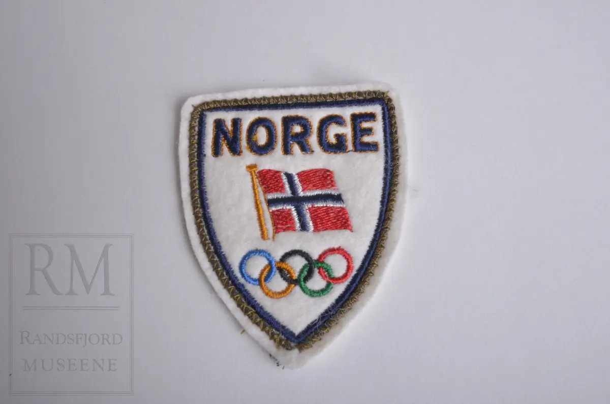 Jakkemerke/strykemerke. "Norge" med Norsk flagg og OL ringene.