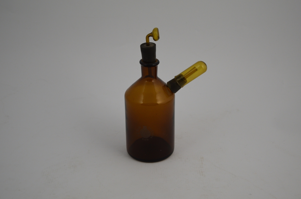 Brun glassflaske, 500 ml. To åpninger, to gummipropper og rør. Ble brukt ved produksjon av øyedråper.