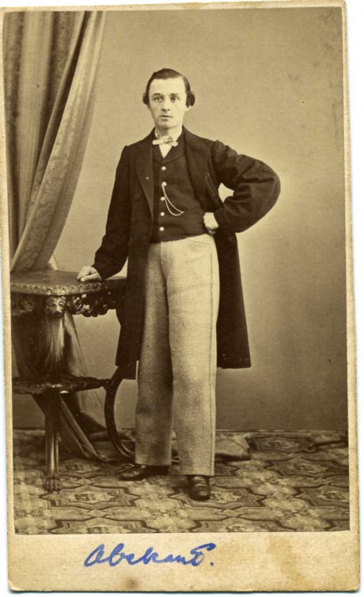Kabinettsfotografi: okänd ung man i ljusa byxor, rock, väst och kravatt, stående vid ett bord.