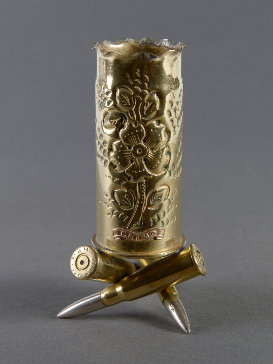 En liten sylinderformet vase i messing, stående på tre geværhylser. På forsiden er det en inngravert blomst og teksten REIMS.