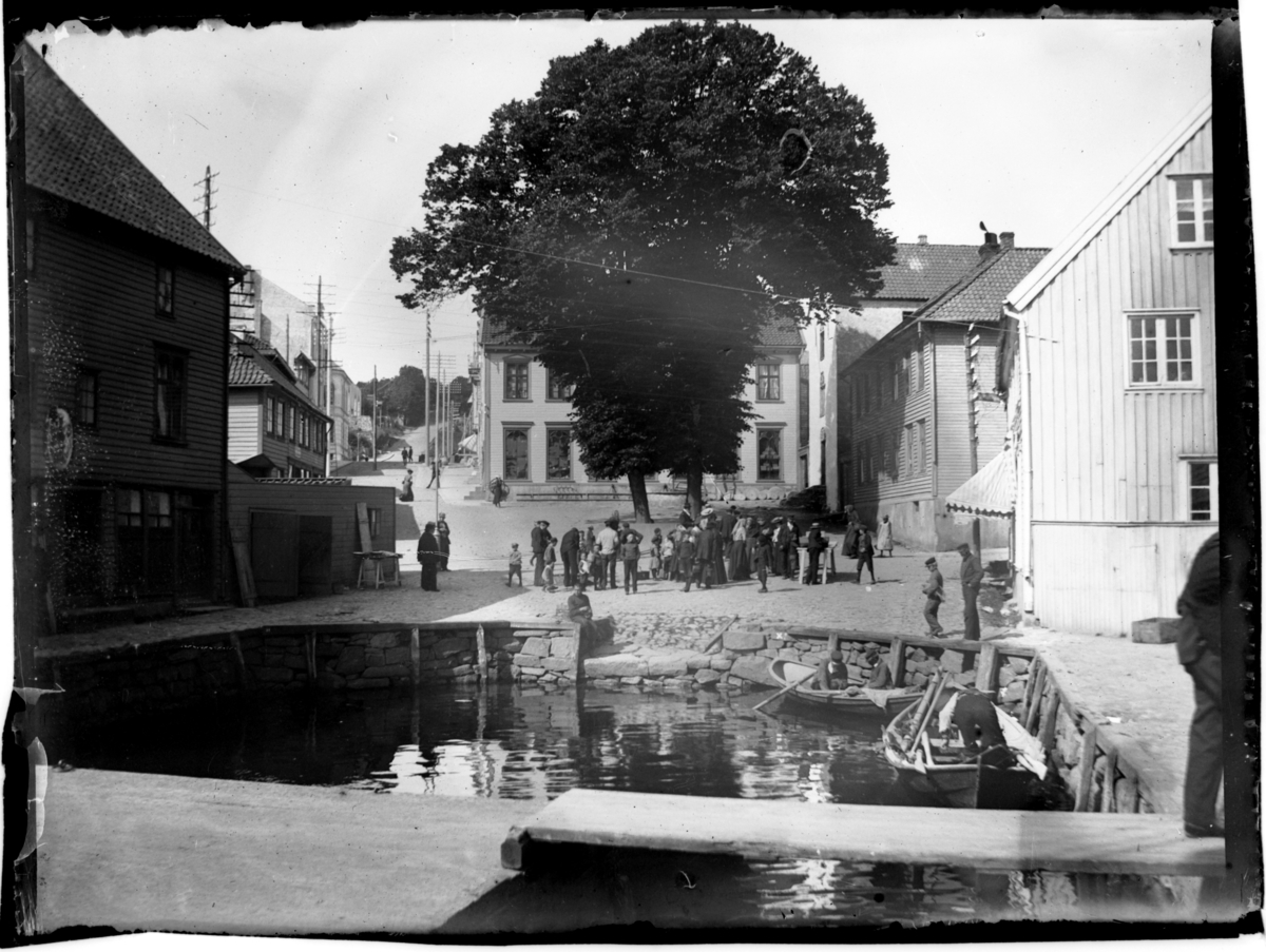 Farsund torv med småbåthavnen i forgrunnen. En gruppe mennesker er samlet på torvet. Fotografert 1903.