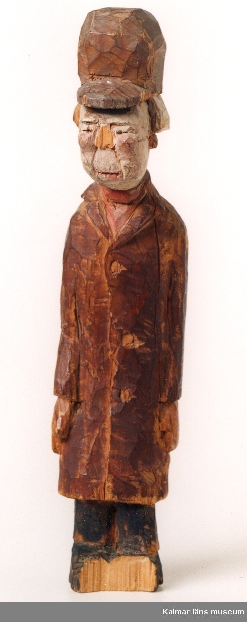 KLM 39255:18. Skulptur, av trä, bemålad. Gubbe i lång, brun rock och hög mössa med skärm. Defekt, näsa och fötter avslagna, samt torrspricka genom överkropp.