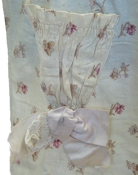 Midjeförkläde av mönstrat bomullstyg i vitt med blommönster. Förklädet är rynkat i midjan, utan linning samt har rosa sidenband till knytband. Nertill avslutas förklädet med en maskintillverkad vit spets. Förklädet har en ficka med en rosa sidenrosett fastydd på utsidan.