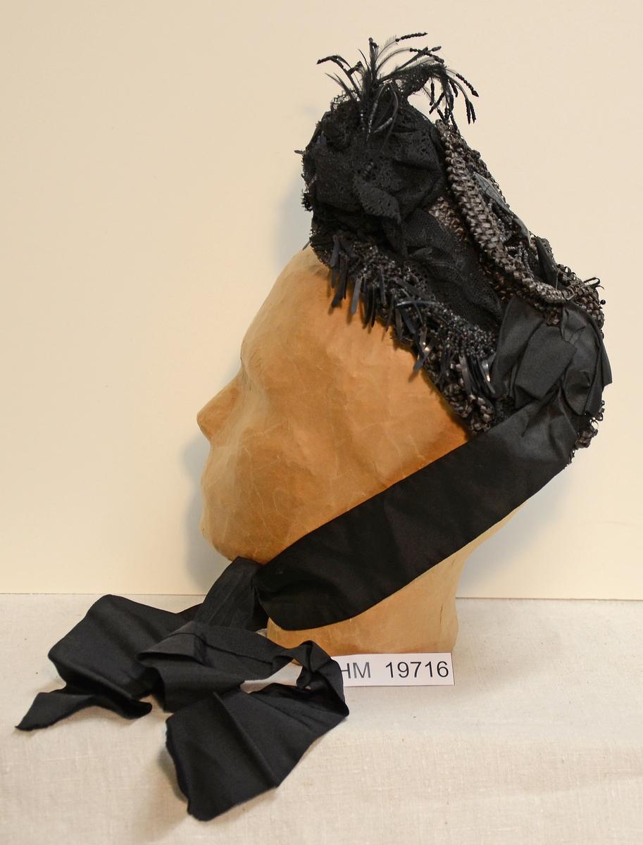 Svart kapotthatt på stomme av ståltråd, prydd med bastflätor, tyllspets mm. Hatten har knytband av svart sidenrips och svart bomullsfoder.