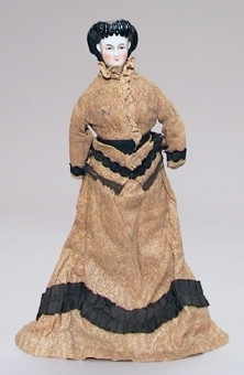 Docka med porslinshuvud med kläder i 1870-talets stil. Beige klänning med rysch och band i brunt. Underkjol av beige brodyr.