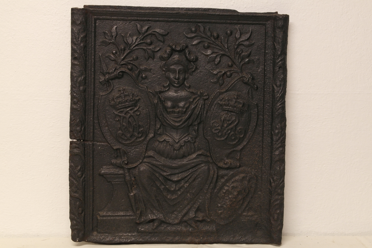 Ovnsplate i støpt jern. Motivet er en brynjekledd, sittende kvinneskikkelse (Minerva) som er flankert av to kronede monogrammer (Kong Christian VI - 1730-1746 - og dronning Sophie Magdalene)