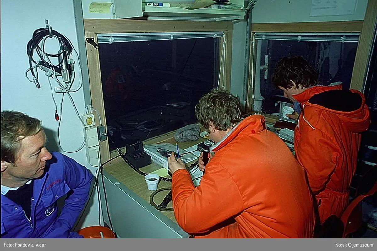 To menn ikledd orange kjeledresser studerer kart i et rom for dykkerkontroll (?)