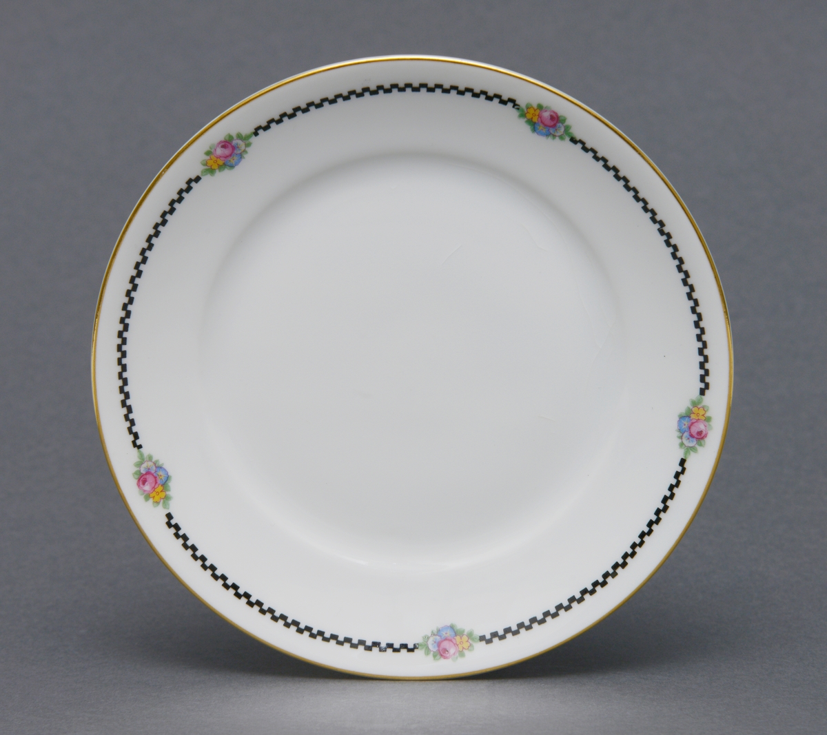Sirkulær tallerken i porselen med glasur. Dekorert med en gullforgylt ring rundt kanten og en svart geometrisk bord med blomstermotiv innimellom borden.