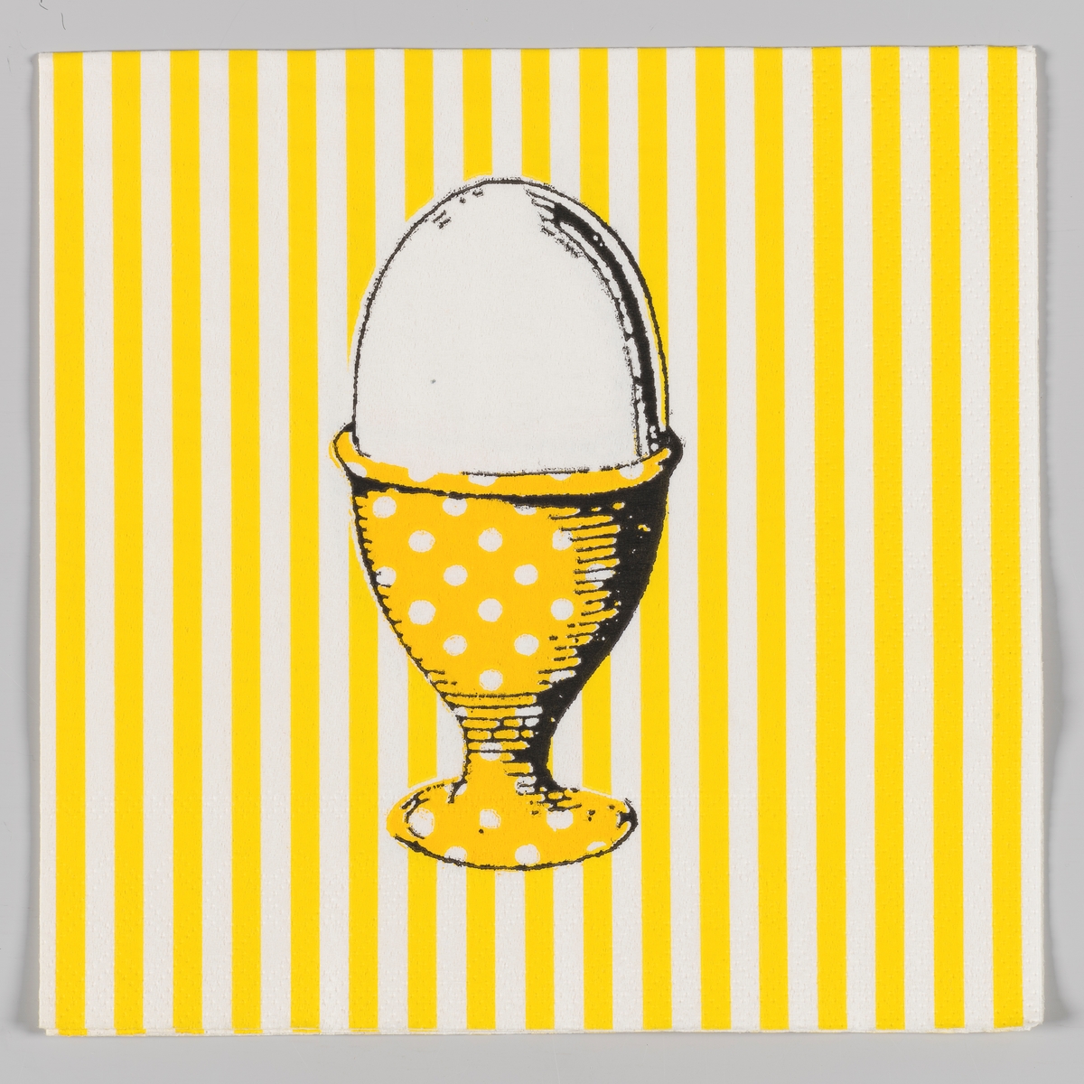 Et egg i et eggebeger utsmykket med hvite prikker på en gul og hvit stripet bakgrunn.