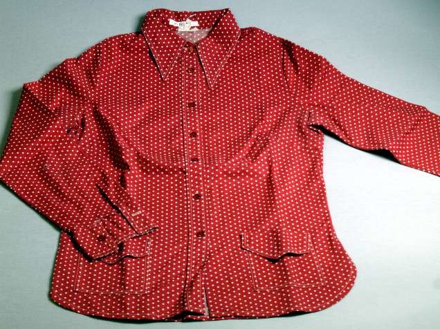 Blus, s.k. utanpåskjorta, röd- och vitprickig av bomull. Påsydda fickor med ficklock fram. Röda plastknappar. Total längd 70 cm. Ärmlängd 59 cm. Axelbredd 36 cm.