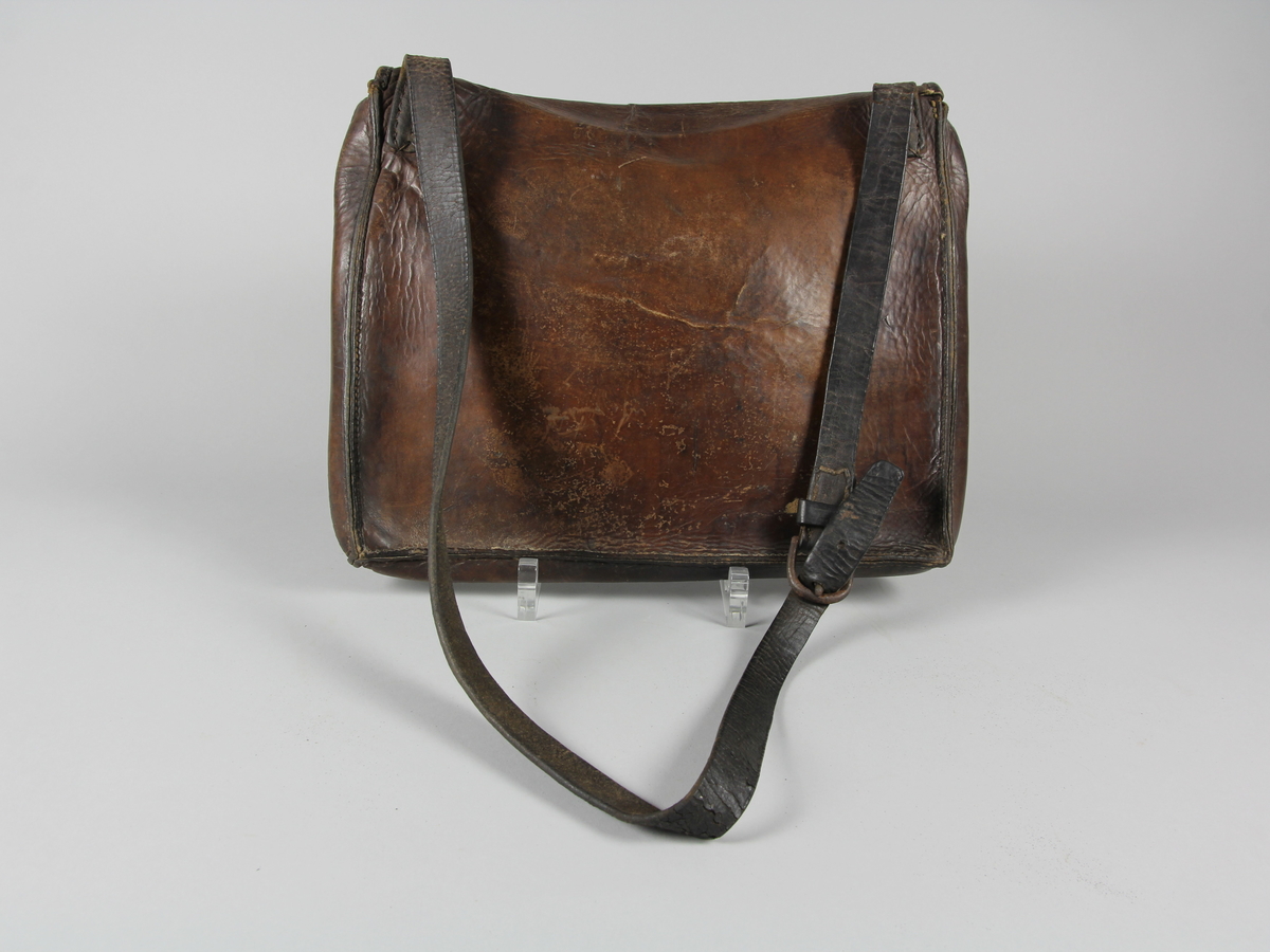 Postväska, rektangulär, av brunt skinn med reglerbar bärrem. Smal klaff/fällock som låses med fyra metallöglor och lång rund metallten.