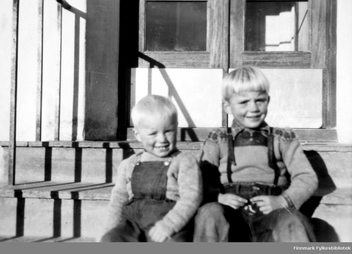 Tor og Kjell Klemetsen. Begge gutter fotografert sittende på trapp med dobbel inngangsdør i bakgrunnen. Gutten til venstre i snekkerbukse/selebukse og gutten til høyre i bukse med bukseseler. Begge har ullgenser.  Familiealbum tilhørende familien Klemetsen. Utlånt av Trygve Klemetsen. Periode: 1930-1960.