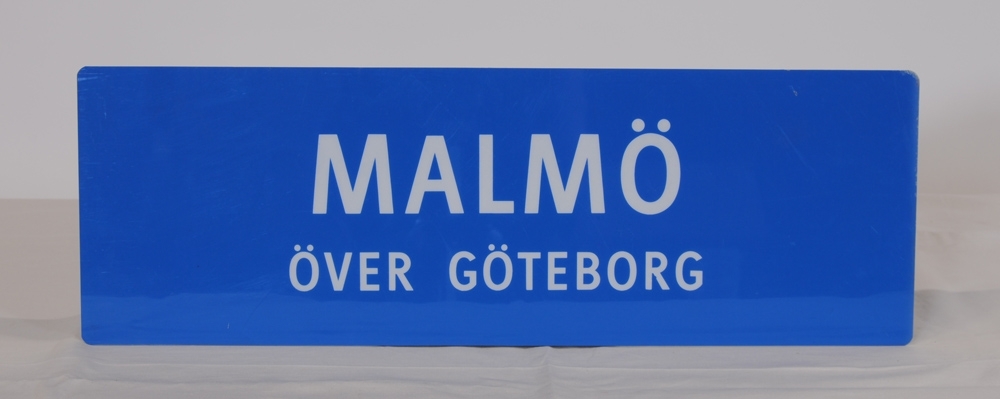 Destinationsskylt av ljusblå plast med texten "MALMÖ ÖVER GÖTEBORG" i vitt. Spegelvänd text på baksidan.