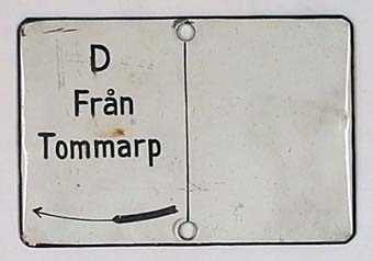 Rektangulär skylt av emaljerat plåt med svart text på vit botten. Skylten är delad i två fält med ett smalt vertikalt streck, med text enbart på vänster sida:
"D
Från
Tommarp".
