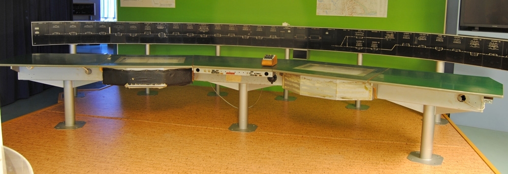Del till skrivbord, nummer 5 från höger. På stolpliknande ben.

Skrivbord till fjärrblockeringscentral. Skrivbordet består av fem ihopkopplade sektioner. Sidorna på mittendelen är vinklade så att skrivbordets sidor är vinklade inåt. Samtliga sektioner har en bordsskiva av trä med en tunn grön plastskiva. Runt hela skrivbordets bordsskiva sitter en grå rundad plastlist. Skivorna sitter fast med gångjärn på baksidan av skrivbordet och går att öppna som en lucka. Innanför sektionerna på sidorna och i mitten (:1, :3 och :5), dvs sektionerna på ben, finns utrymme för sladdar. Sektionerna på sidorna har ett sladdhål på baksidan, och sektionen i mitten har två, ett vid vardera kant samt ett telefonjack. Innanför de två sektionera utan ben (:2 och :4) finns varsin skrivare. Benen är stolpliknande och sitter på plattor som skruvas fast i golvet med fyra skruvar. De två sektionerna utan ben är hopkopplade mellan två sektioner med ben. Dessa platser är avsedda för att sitta och arbeta vid med tanke på benutrymmet. Dessa bordsskivor är utrustade med ett fönster. Sargen runt skrivaren är stoppad och klädd med fuskläder på ena sidan. På andra sidan finns limrester och där har det sannolikt sett ut på samma sätt. På sargen av mittendelens bordsskiva (:3) sitter två lås samt några knappar och spakar. På sidorna är det varsitt hål. Längre ned, ovanför benet, sitter en lapp som beskriver reglagen inklusive de tomma hålen fr.v: 1. Nödstopp Kmb-Sbk, m2. Nyckel För manövrering av NSB , 3. Nyckel För manövrering av NSB, 4. Manöversändare Reserv, 5. Indikeringsmott Reserv, 6. Knappsats, 7. Avstängning av akustiskt larm (ras och skred), 8. Knappsats. Norr, söder eller båda linjerna, 9. Frånskiljare, tänd el släckt., 10. Manöversändare Reserv, 11. Indikeringsmott Reserv, 12. Nödstopp Kra-Håk. Rsi-Svv. Till höger om lappen sitter en apparat som ser ut att vara en timer. Apparaten är märkt "POWERSTAT" och "THE SUPERIOR ELECTRIC CO BRISTOL. CONN. USA". Några av reglagen är dessutom märkta med plastremsor med text. Vid de två reglagen till höger om nyckelhålen står det: FRÅN, TILL, LINJE 1 MS RES rep LINJE IM RES. Vid reglaget i mitten står det med en röd plastremsa: AVSTÄNGNING AV AKUSTISKT LARM FÖR RAS OCH SKRED. Vid de tre knapparna till höger står det: SLÄCKT, TÄND, ELA. BEL., L3, MS, L4, L3 och IM. På vänstra delen (:5) sitter ett eluttag och en grön knapp och på vänstra delen (:1) sitter en lampa och ett lås.