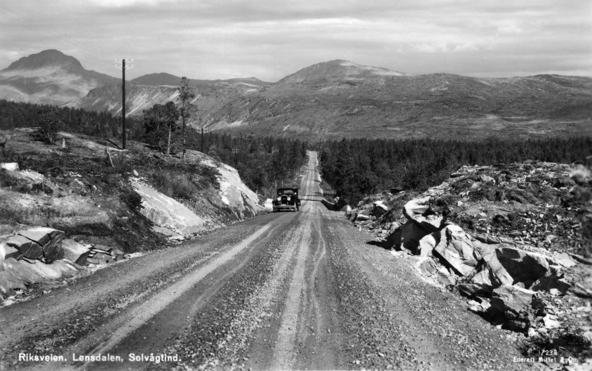 Riksvegen gjennom Lønsdalen (Luonosvagge) i Saltdal kommune i Nordland, slik den så ut i 1930-åra.  Fotografen har stått på et sted hvor vegen går i ei lav skjæring over et lite bergparti.  En bil er på veg opp mot opptaksstedet.  I bakgrunnen en forholdvis lang rett strekning gjennom glissen skog.  Over skogen troner en fjellrygg, muligens Tjørnfjellet (Girnno).  Helt til venstre rager Solvågtind mot himmelen.  