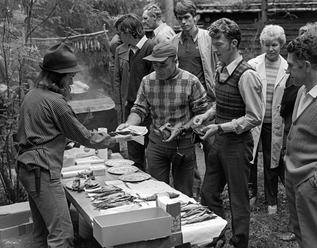 Salg av varmrøykt kvitfisk under De nordiske jakt- og fiskedager på Norsk Skogbruksmuseum i 1973.  Fiskerøykinga foregikk i et nylagd anlegg, med muligheter for både kald- og varmrøyking, i friluftsmuseet på Prestøya.  Her ser vi Gunhild Eknæs, kledd i busserull og med hatt på hodet, som serverte fisk med flatbrød og smør på papptallerkener fra et bord, mens en liten kundeflokk ventet.  I bakgrunnen skimter vi røykeanlegget.  I 1973 var røykeanlegget for fisk på vestsida av Prestøya nytt.  Man kunne vise både kaldrøyking og varmrøyking, men det var den sistnevnte metoden som var raskest, og dermed best egnet for dette arrangementet.  Varmrøyking er en tilberedningsmåte for fisk på linje med koking og steking.  Den forholdsvis svale og kortvarige røykinga har begrenset effekt, og varmrøykt fisk har følgelig ikke like lang holdbarhet som kaldrøykt.  Nytilberedt varmrøykt fisk er imidlertid delikat og velsmakende.  Varmrøykingsanlegg lages gjerne på en bakkekam, med en forholdsvis kort røykkanal fra et punkt nede i bakkeskråningen opp mot ei tønne eller kasse, der fisken henges eller legges, og hvor røyken samles.  Ved Skogmuseets røykeanlegg på Prestøya er det et kasseformet røykkammer med skuffer eller brett med nettingbotn, som fisken legges på.  Kassa dekkes med et strieklede.  Etter innledende fyring med ved, som etterlater seg glør i den nedre delen av røykkanalen, fyres det blant annet med einerbar, som gir fin røyk.  Temperaturen inne i røykekammeret bør være mellom 60 og 80 grader.  Det tar fra 45 minutter til to timer å få fisken serveringsklar, avhengig av fiskens størrelse, fyringa og av hvilket smakspreg som ønskes. 
