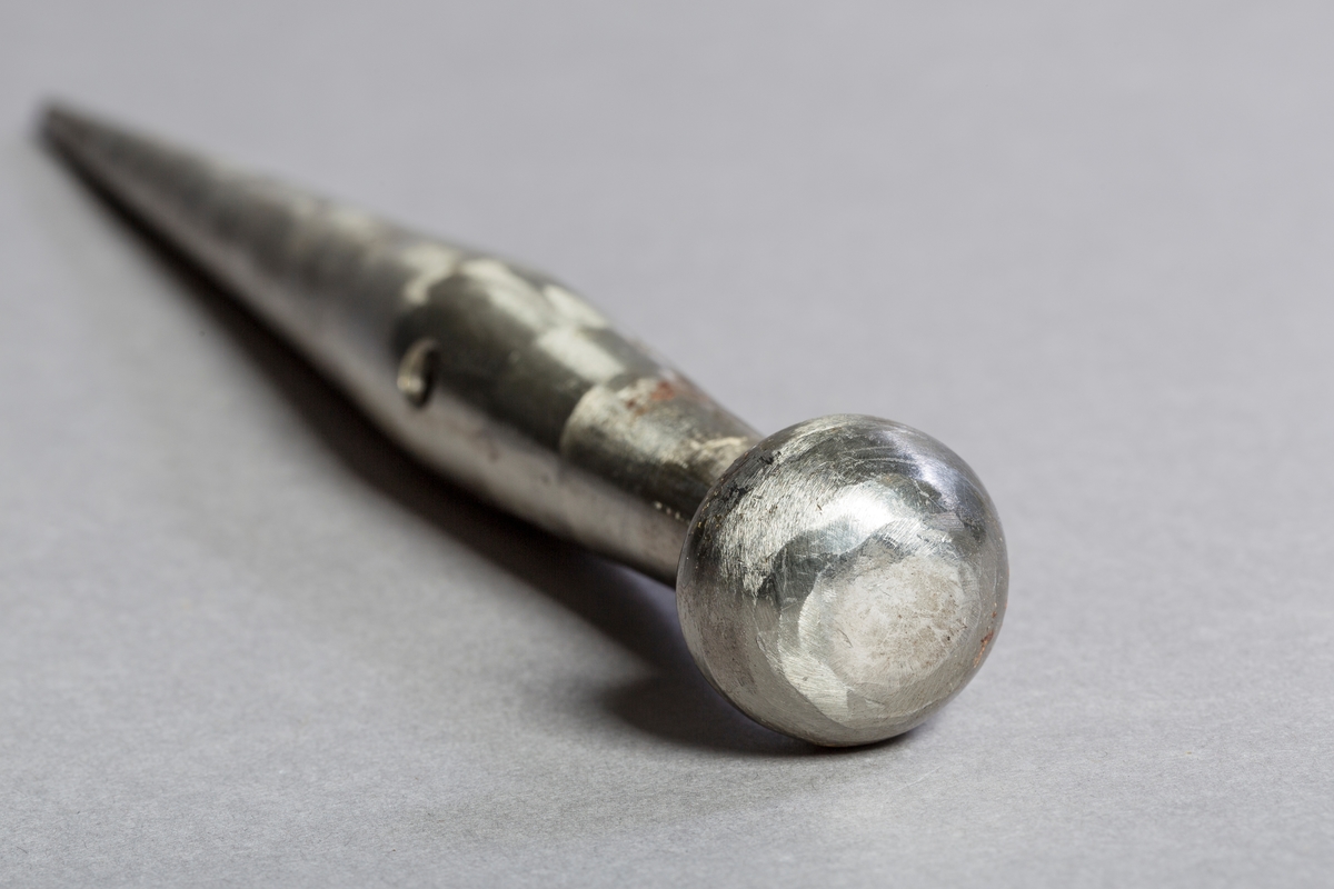 En avlang sylinderformet stang i metall som er spiss i den ene enden og har en halvkuleformet hode i den andre enden. Det er et gjennomgående hull på det tykkeste partiet midt på stangen.