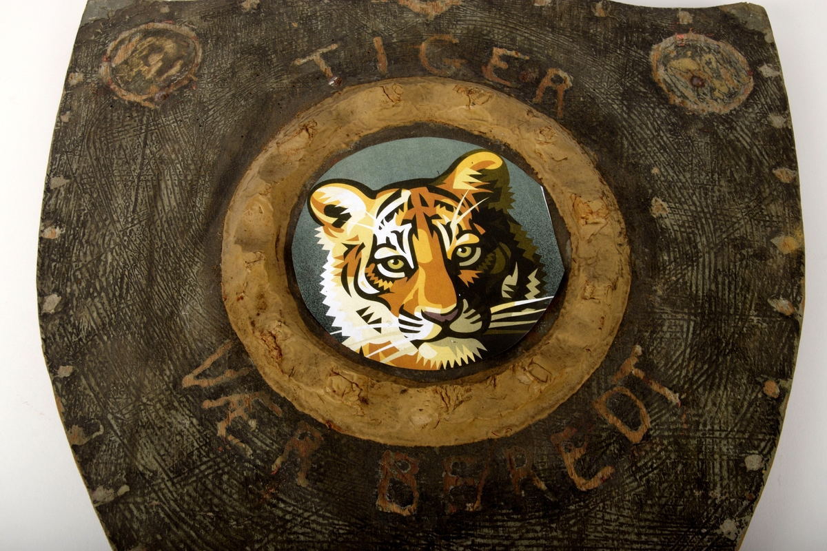 Treskjold med dekorativ utskjæring i toppen. Midt på skjoldet er en forhøyning hvor et tigerbilde er pålimt - logo for "Tigerpatruljen". 

På baksiden er to trestykker for støtte, samt et tau til oppheng.