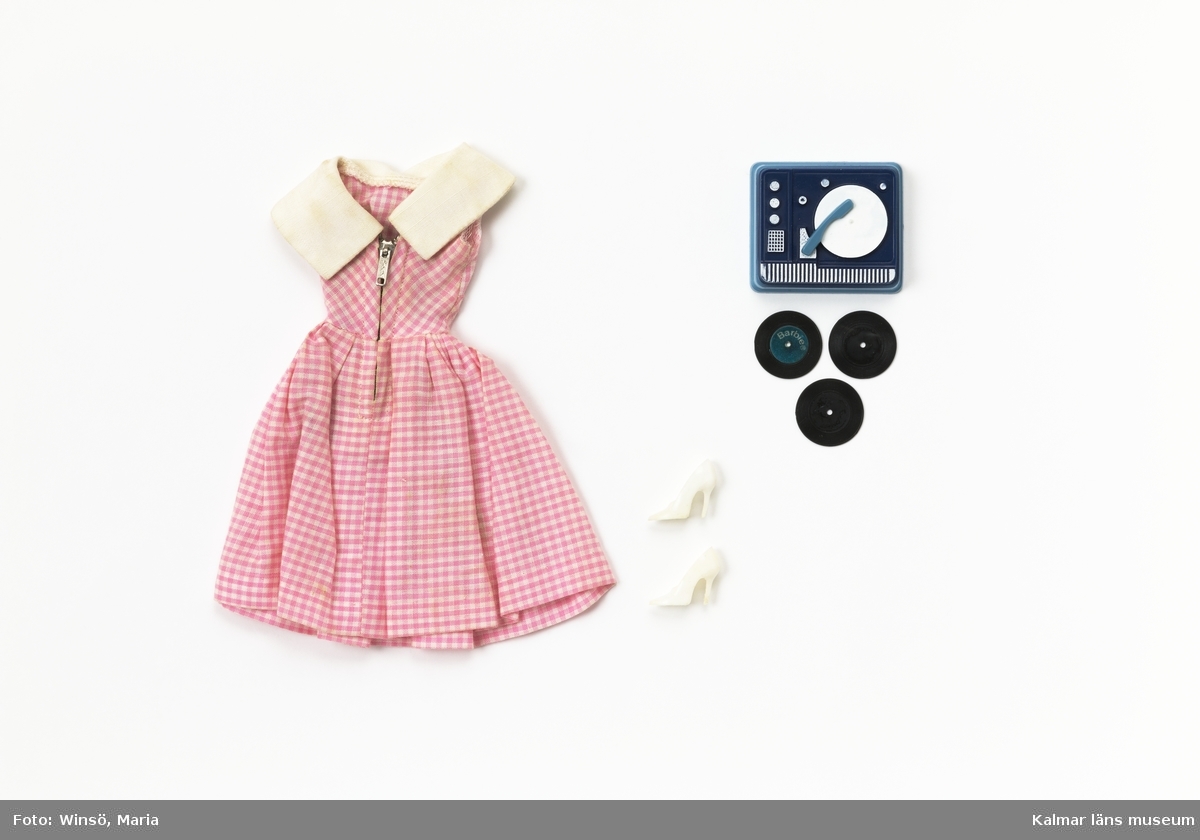 KLM45643:19 Dockkläder. Klänning, skor, grammofon, grammofonskivor. Set med klänning i rutigt tyg i rosa och vitt med ett vit ok broderat med rosor, vita högklackade plastskor, tre skivor av svart plast och en skivspelare av blå och vit plast. Klänningen är märkt med Mattels etikett. Setet finns i häftet ”Barbie och hennes vänner – med många nya kläder” från Brio och heter ”626 Grammofonmusik och dans”. Setet är för Barbie.