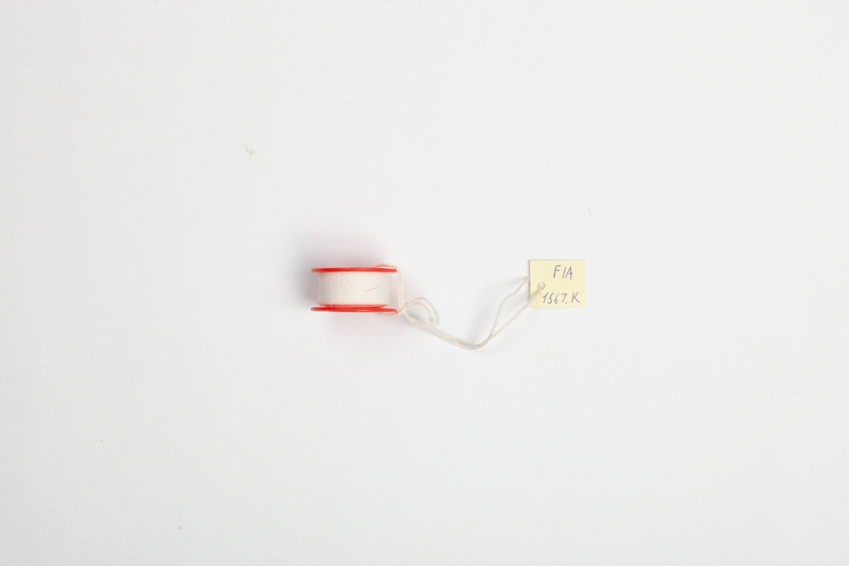 En liten rull med hvit bandasjeteip. Teipen er anbrakt på en rød rund plastramme med et rundt hull.