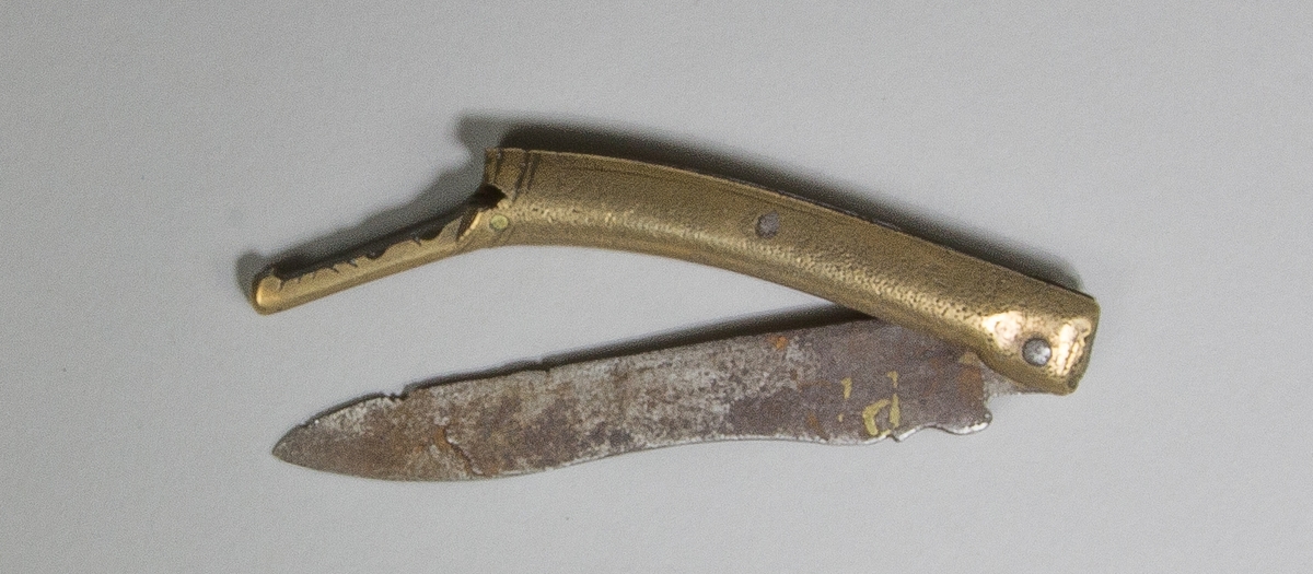 Gästabudskniv, av stål, med ledat handtag av järn och mässing. Handtaget med graverad dekor, bakre delen i form av underkäken på ett drakhuvud. Längst bak på handtaget ett spår för en mindre gaffel.