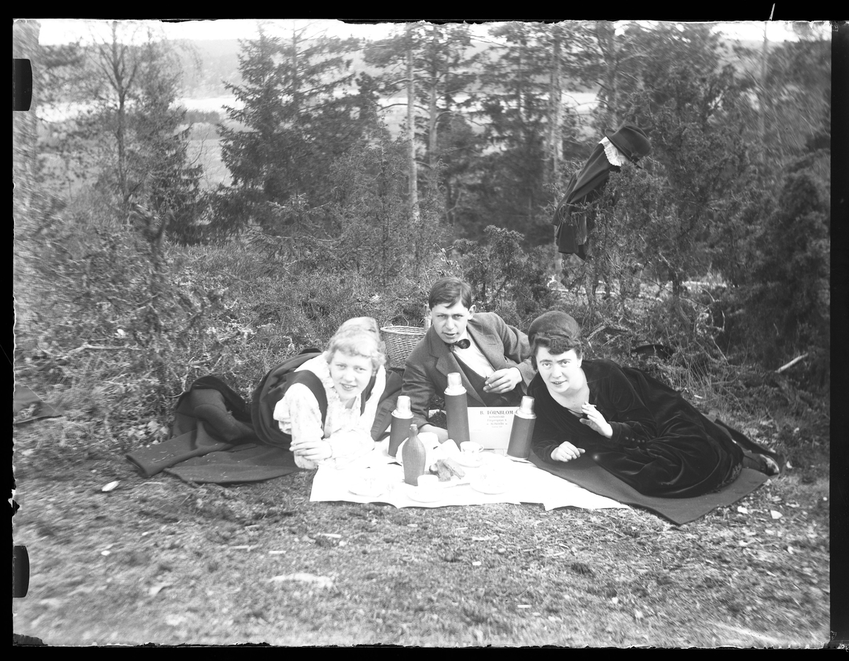 Signe Thorsell, Hugo Larsson och Eva R ligger på filtar i gräset, röker och fikar. Framför dem är termosar, koppar och en flaska uppdukade. Fotot taget på Kristi himmelsfärdsdagen.