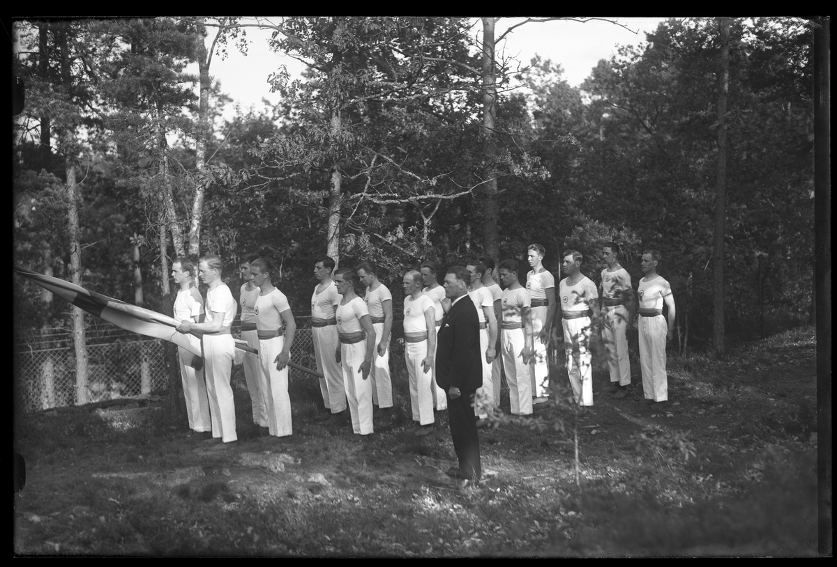 Gruppbild på Alingås Gymnastikförenings manliga trupp. Gymnasterna har vita dräkter och i mitten står en man i kostym. Den ena mannen håller i en fana. I fotografens egna anteckningar står det "A.G.F.s uppvisningar i Parken".
