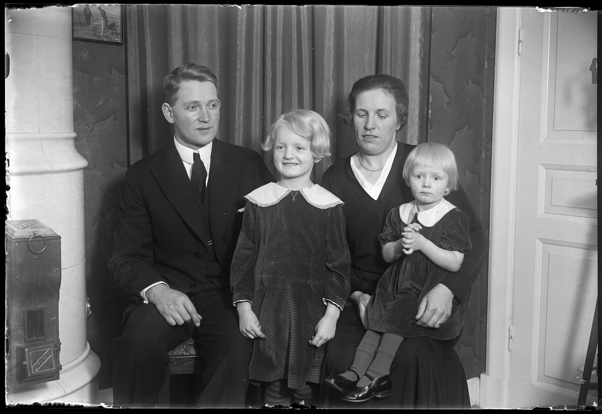 Familjeporträtt på familjen Dalén med mamma Tullie, pappa Carl samt barnen Ann-Marie och Margit. Familjen sitter vid en kakelugn och alla bär mörka kläder. I fotografens egna anteckningar står det "Carl Dahlén med familj".