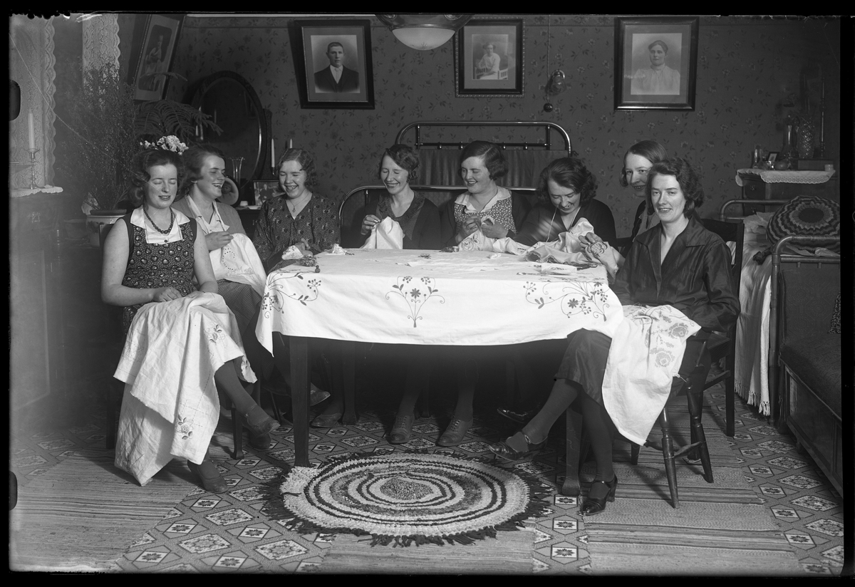 8 glada kvinnor sitter runt ett bord och handarbetar. I fotografens anteckningar står det "Symöte hos Gust. Johansson".