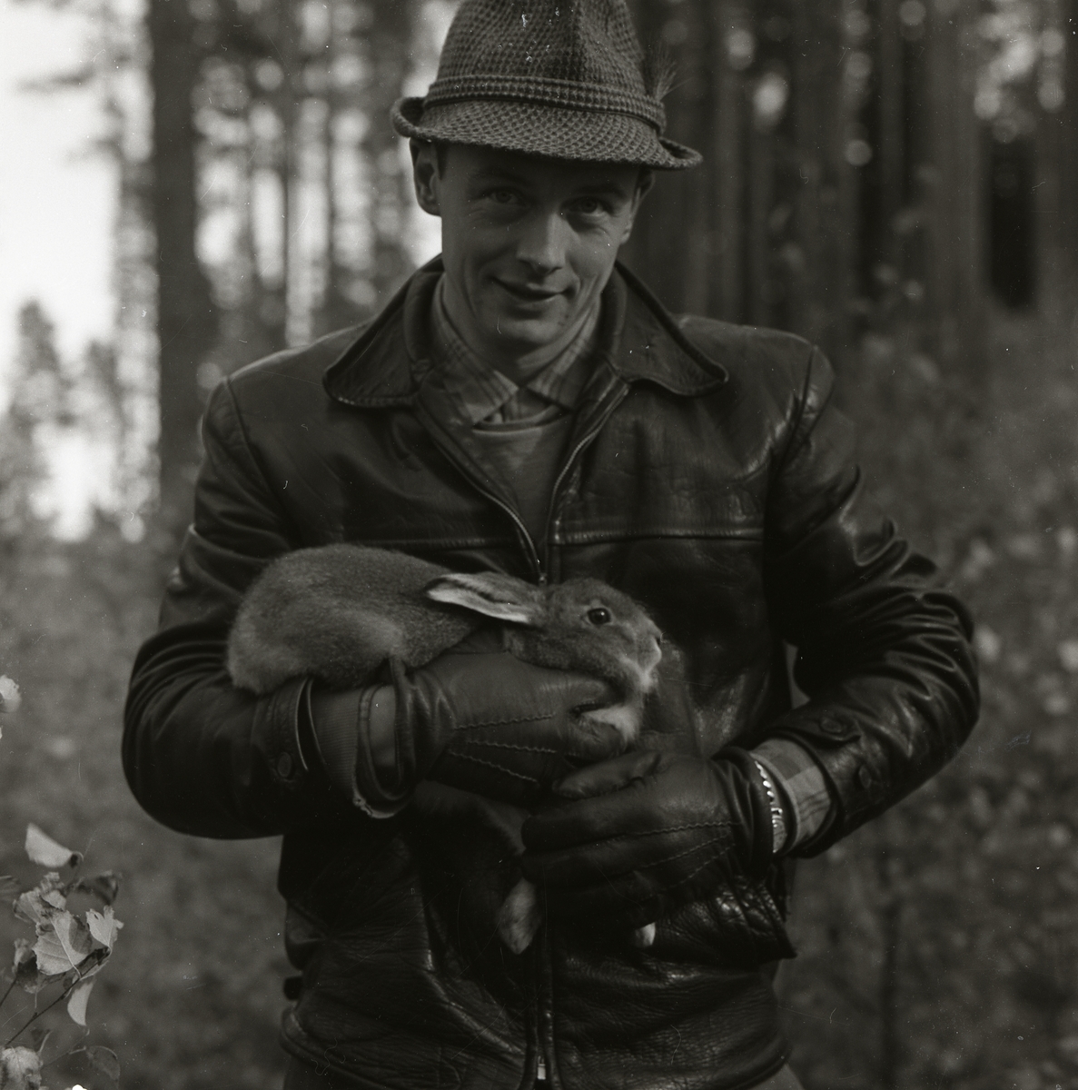 En man iförd hatt och jacka står i skogen med en hare i famnen på Storön, 6 oktober 1957.