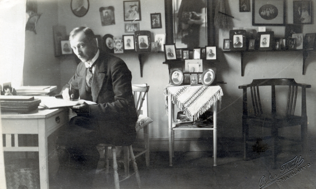 Oskar Träff sitter vid ett skrivbord placerat vid fönstret i ett rum. Längs väggen står ett flertal inramade porträttfotografier. I mitten hänger en spegel i vilken man kan se fotografen och kameran.
