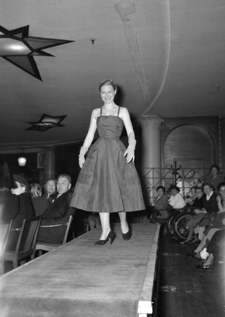 En kvinne på podiet iført kjole med stropper. Fra en motevisning på Steen & Strøm fra 7. mai 1954..