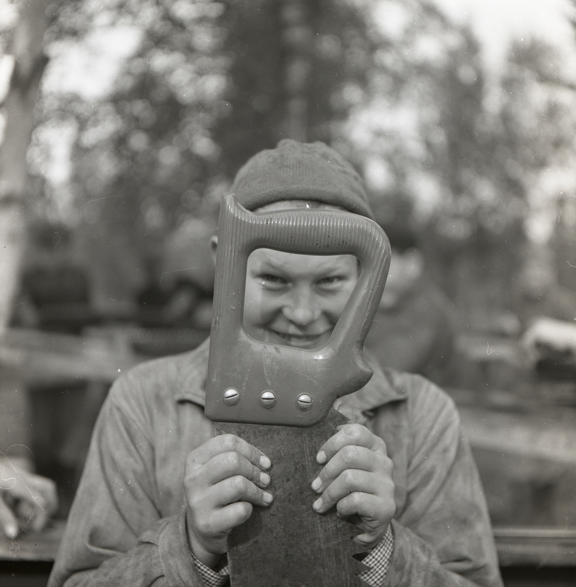 En pojke håller upp en såg framför ansiktet och tittar mot fotografen genom handtaget, Norrvåga 1953.