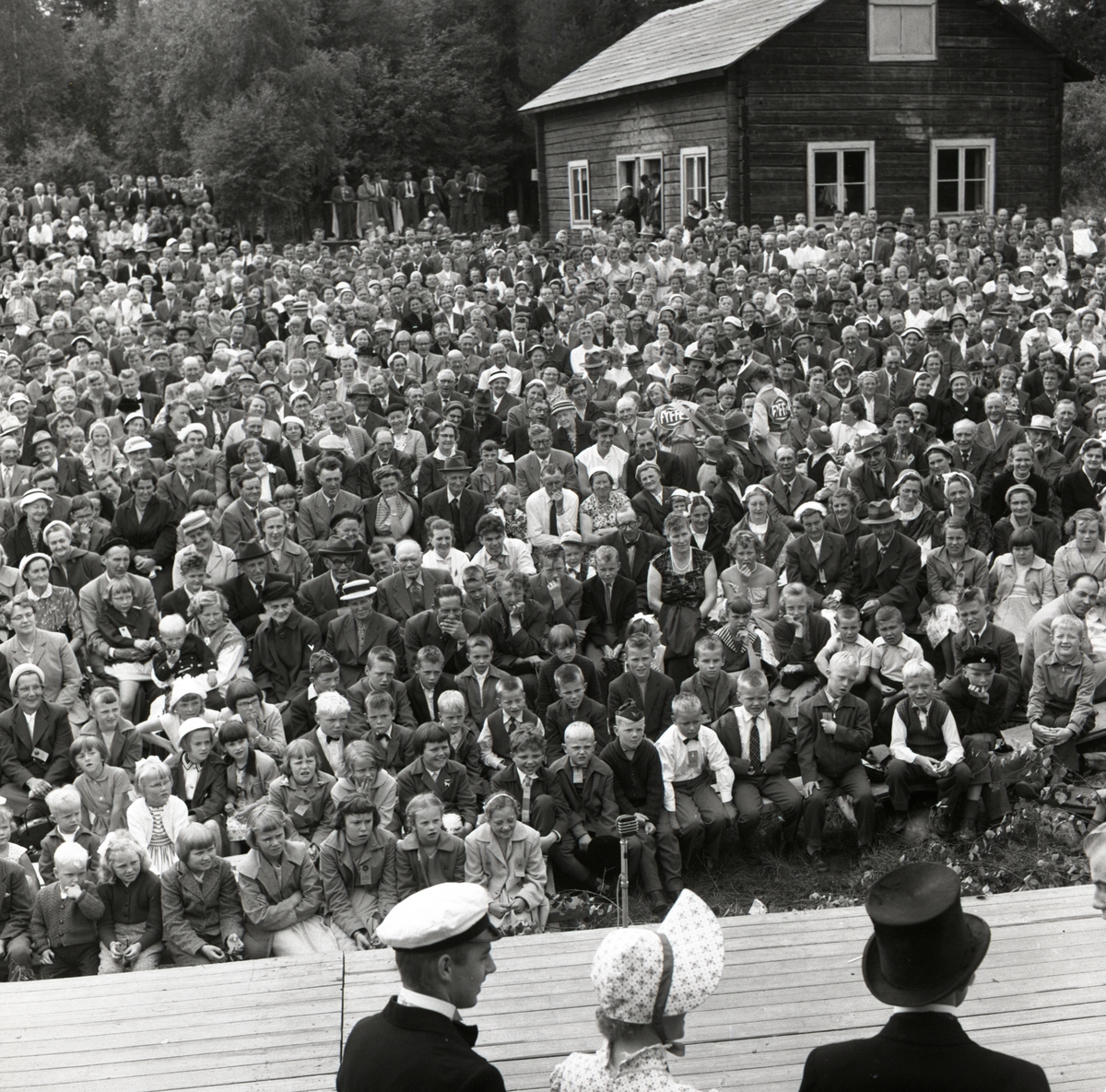 Tre personer iklädda huvudbonader står på scen framför ett publikhav, midsommarafton den 22 juni 1958 i Rengsjö.
