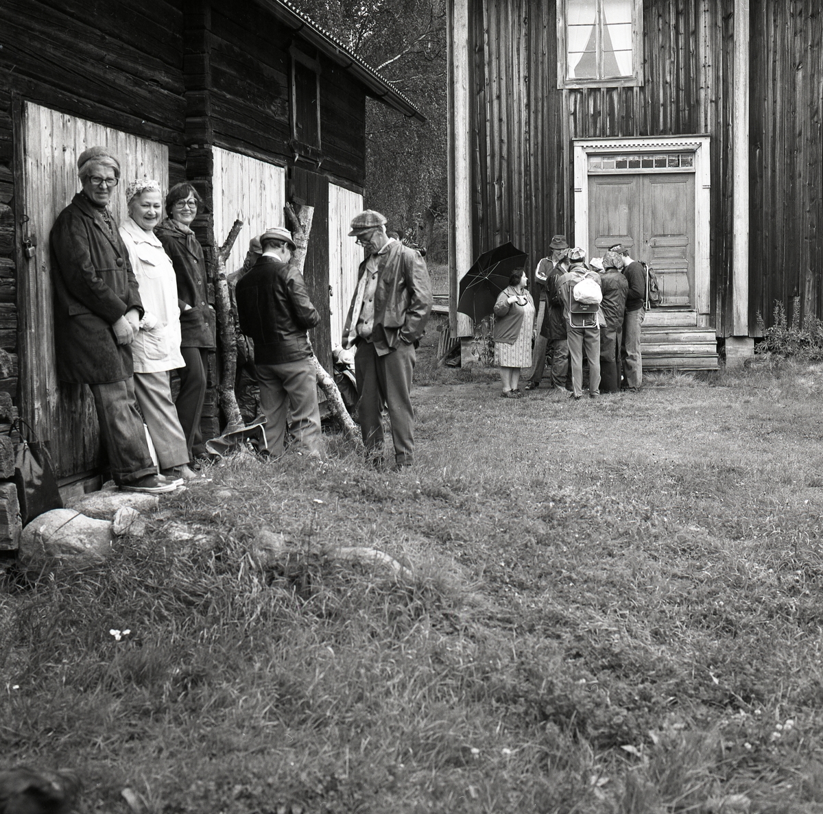 En grupp människor står intill väggarna vid några hus och väntar på invigning av en kyrkstig vid Tannåsen, september 1977.