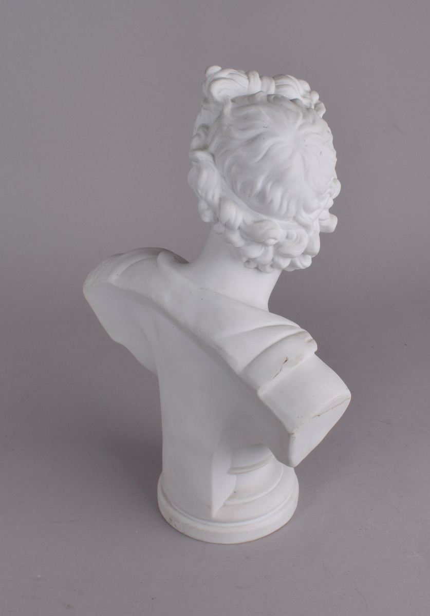Klassisk mannsfigur med håret i sløyfe og draperier rundt skuldrene. Den romerske/greske guden Apollo