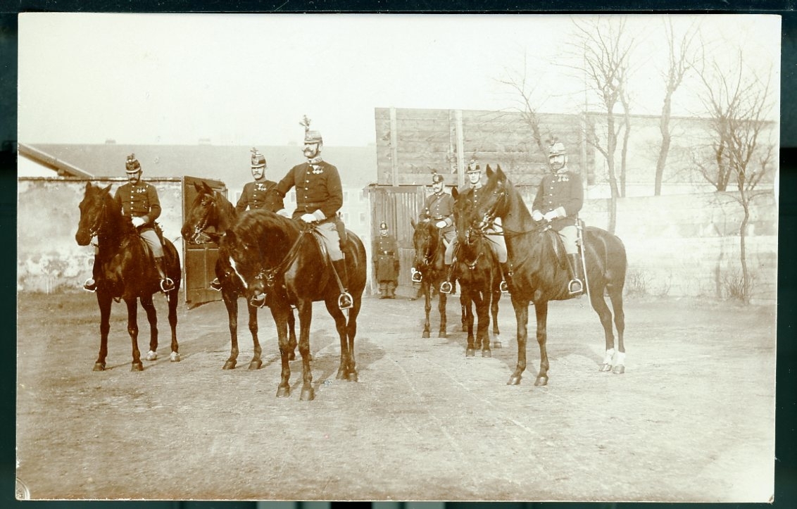 Sex militärer till häst. De står framför en hög mur med port där en soldat står på post.
