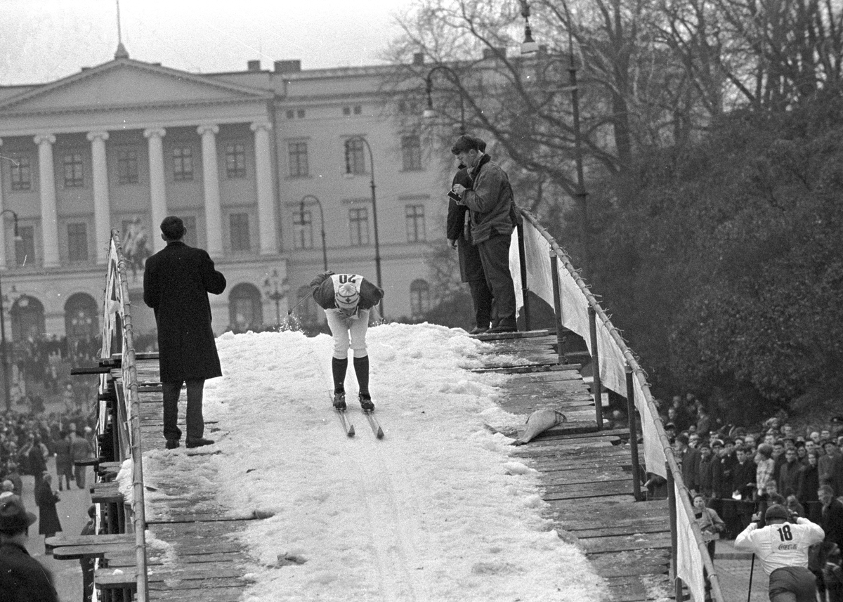 Skiløper på vei over bro, pressefolk står langs løypa, Slottet i bakgrunn, Slottsrennet. Fotografert november 1964.