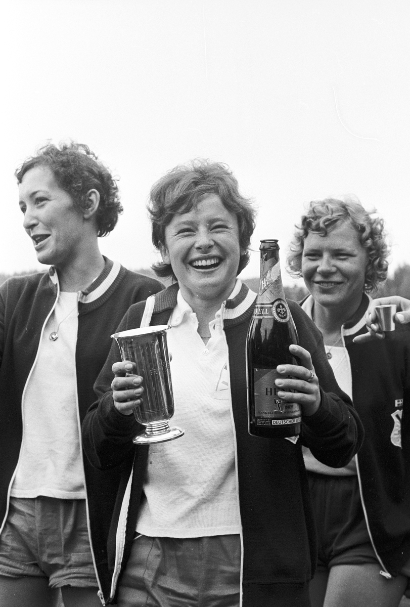 Tre kvinnlige roere, den ene holder en pokal og en flaske sjampanje, NM i roing. Fotografert 1965.