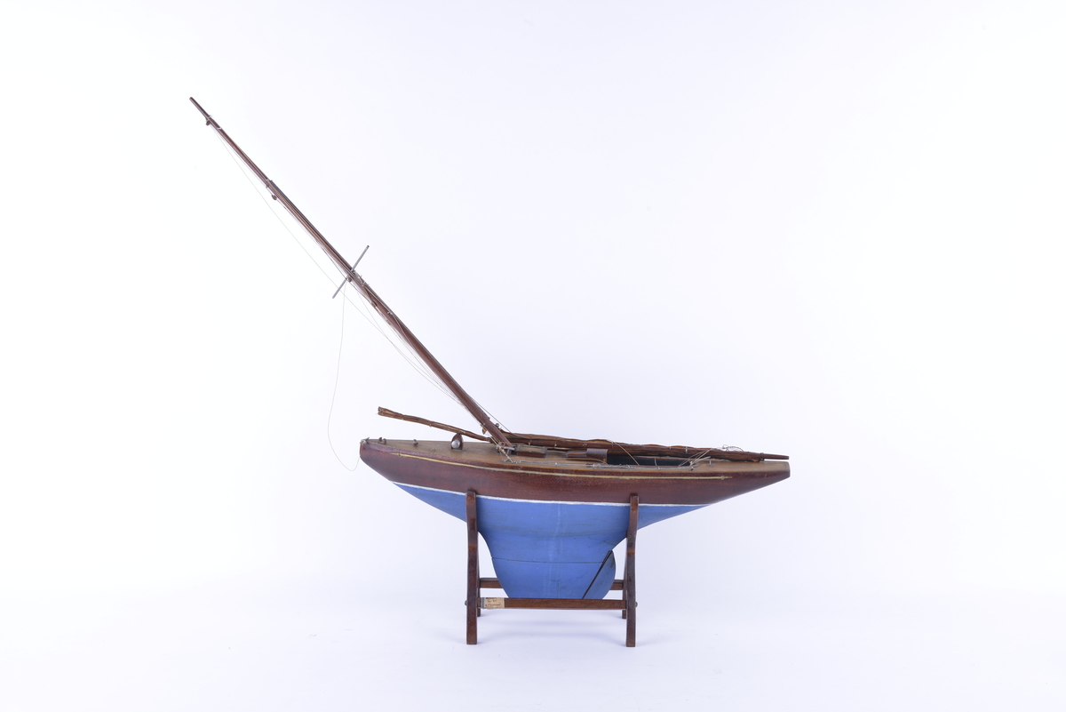 Båtmodell med en hovedmast, samt en mast som er knekt og ligger løst i modellen. Modellen hviler på et stativ av treverk. Ingen seil.
