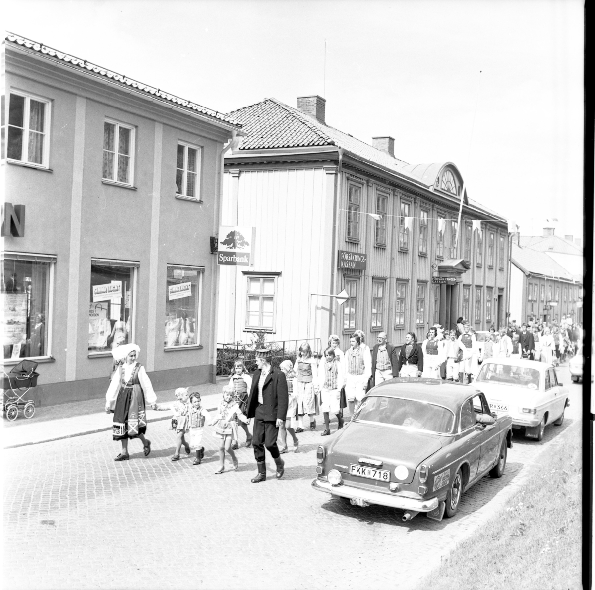Folkdansare marscherar söderut på Brahegatan, i täten en kvinna och en man med tre barn mellan sig. I samband med Grännapolkan på Grännaberget, 1975. Till vänster skymtar Sparbanken.