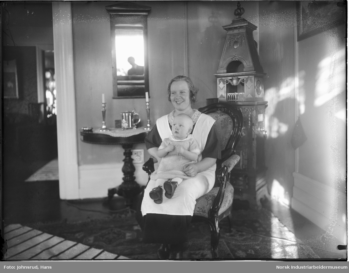Kvinne med barn på fanget sittende i stol i stue. Fotografen synlig i speilet bak kvinnen.