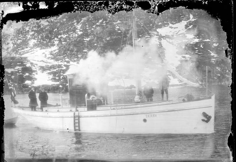 Skandferkutteren "Skarv" fotografert fra Jürgensenkaia mot Øyfjellet. Personer ombord og kutteren spyr opp mye røyk.
