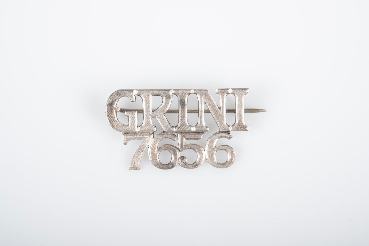 En nål i metall med teksten "Grini 7656". Nål bak som festes i en krok.