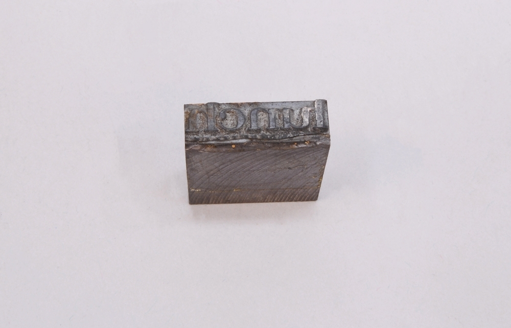 Rektangulär kliché av grå metall, monterad på en grå metallkloss. På klichén finns texten: "lunch", i relief.