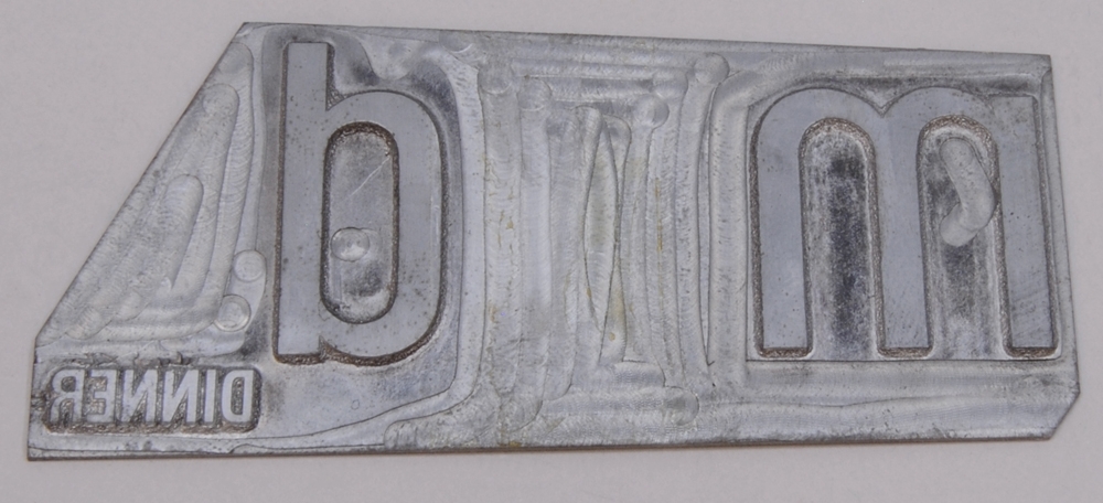 Kliché av silverfärgad metall. Den är rektangulärt formad där det övre vänstra hörnet är beskuret. Från höger finns bokstäverna "m" och "d" i relief. Mellan bokstäverna finns mellanrum. Bakom bokstaven "d" finns texten. "DINNER" i relief.