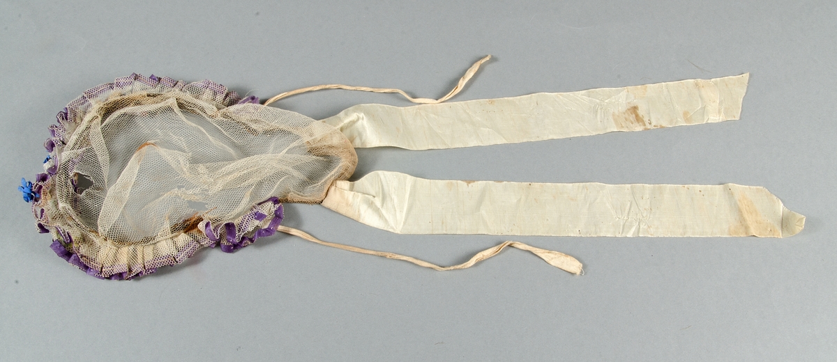 Hårklädsel av vit tyll med två rysch kantade med lila sidenband. Två konstgjorda blå blommor. Två våta taftsidenband. Knytband av vit bomull.